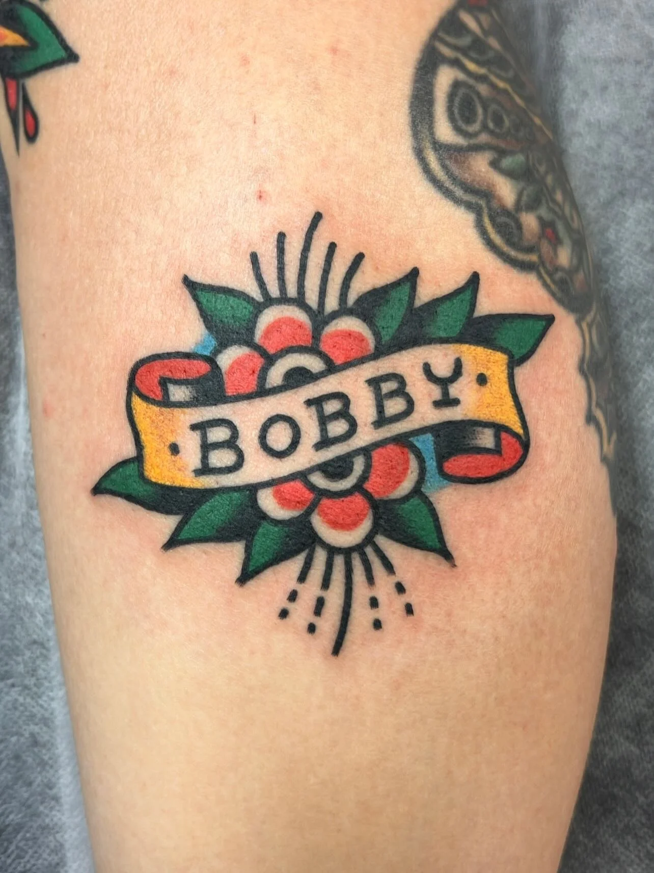 A colorful tattoo features a floral design with a yellow ribbon banner across the center. The banner has the name "BOBBY" written in black uppercase letters. The flower behind the banner has red and white petals with green leaves and radiating black 