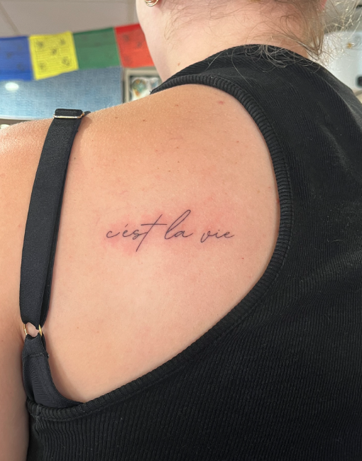 A person's shoulder with a tattoo that says 'c'est la vie' in cursive script.