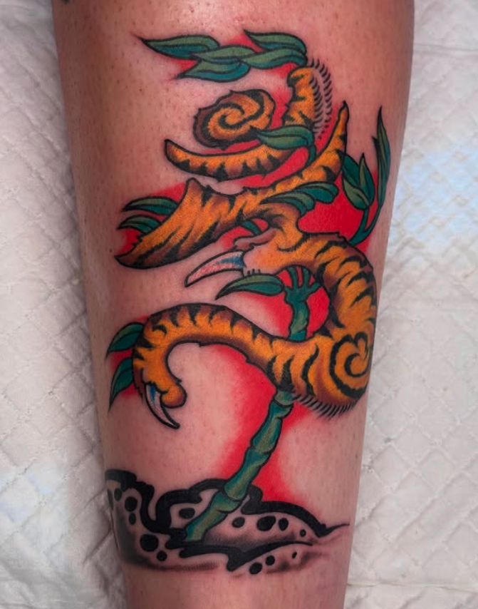 Tattoo of a dragon intertwined with a plant on a person's leg, with a red background and stylized ground at the bottom.