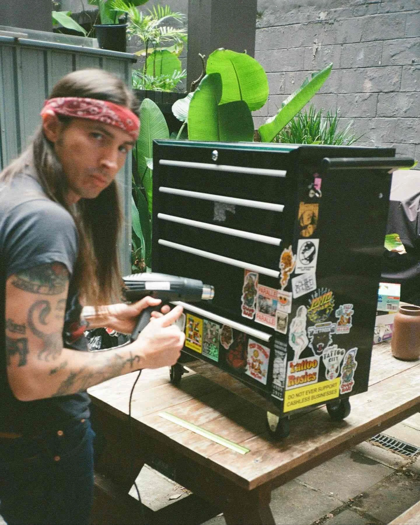 A man with tattoos and a red bandana working on a tattoo machine in front of a decorated tattoo vending machine outdoors.