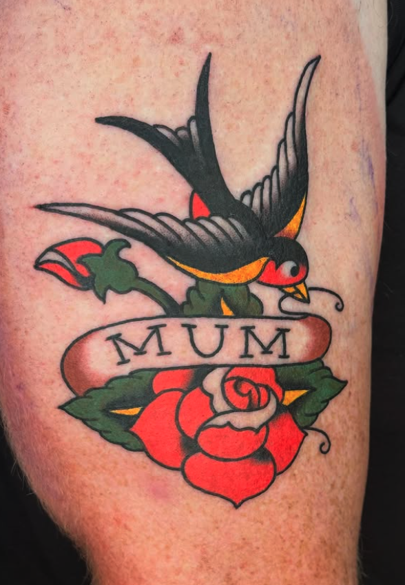 Tattoo of a black swallow bird with red and yellow accents, perched on a red rose with green leaves, and a banner reading 'MUM' in black letters.