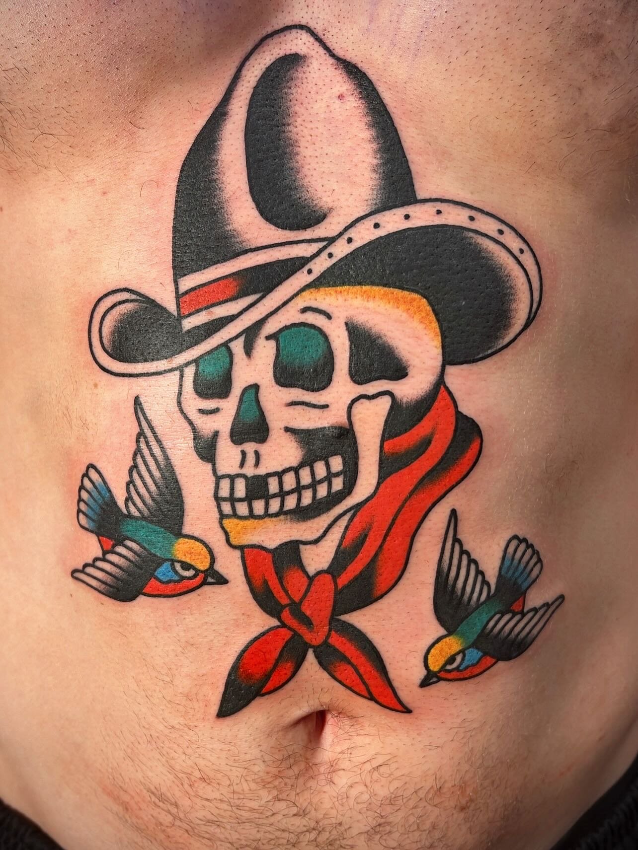 Tattoo of a skull with a sombrero, red bandana, and two colorful birds.