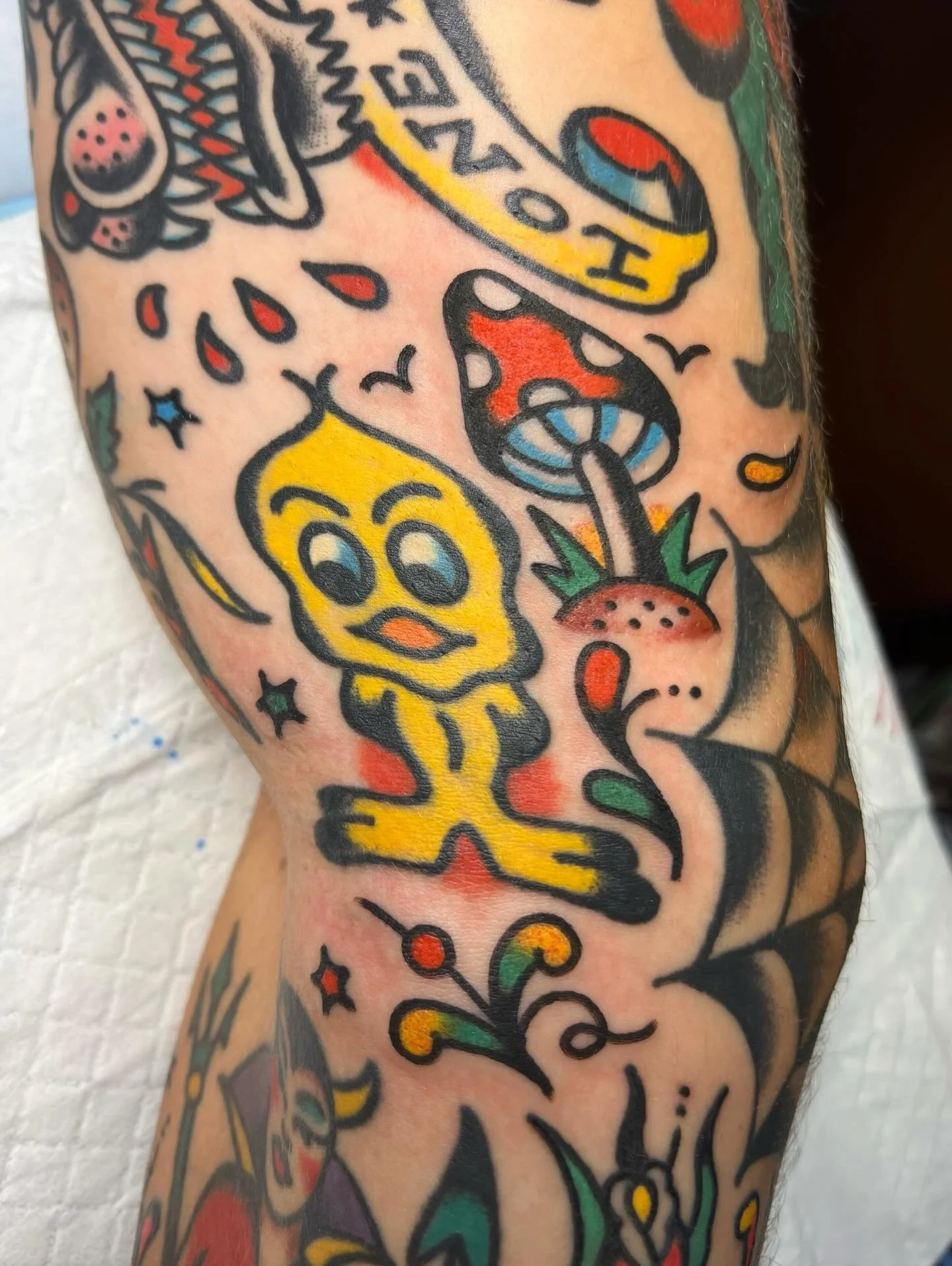 Colorful tattoo on skin featuring a yellow cartoon duckling with big eyes, a red strawberry, a blue mushroom, and various stars and decorative elements.
