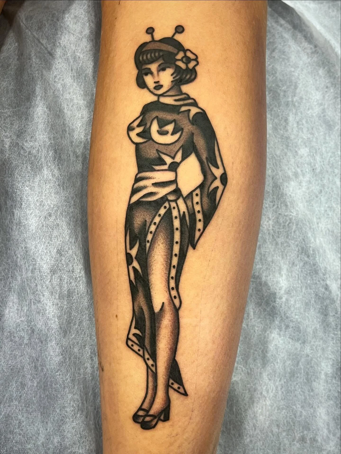 A tattoo of a vintage-style cartoon woman with a bob haircut, antenna headband, and a confident pose, wearing a detailed dress with high heels, on a person's arm.