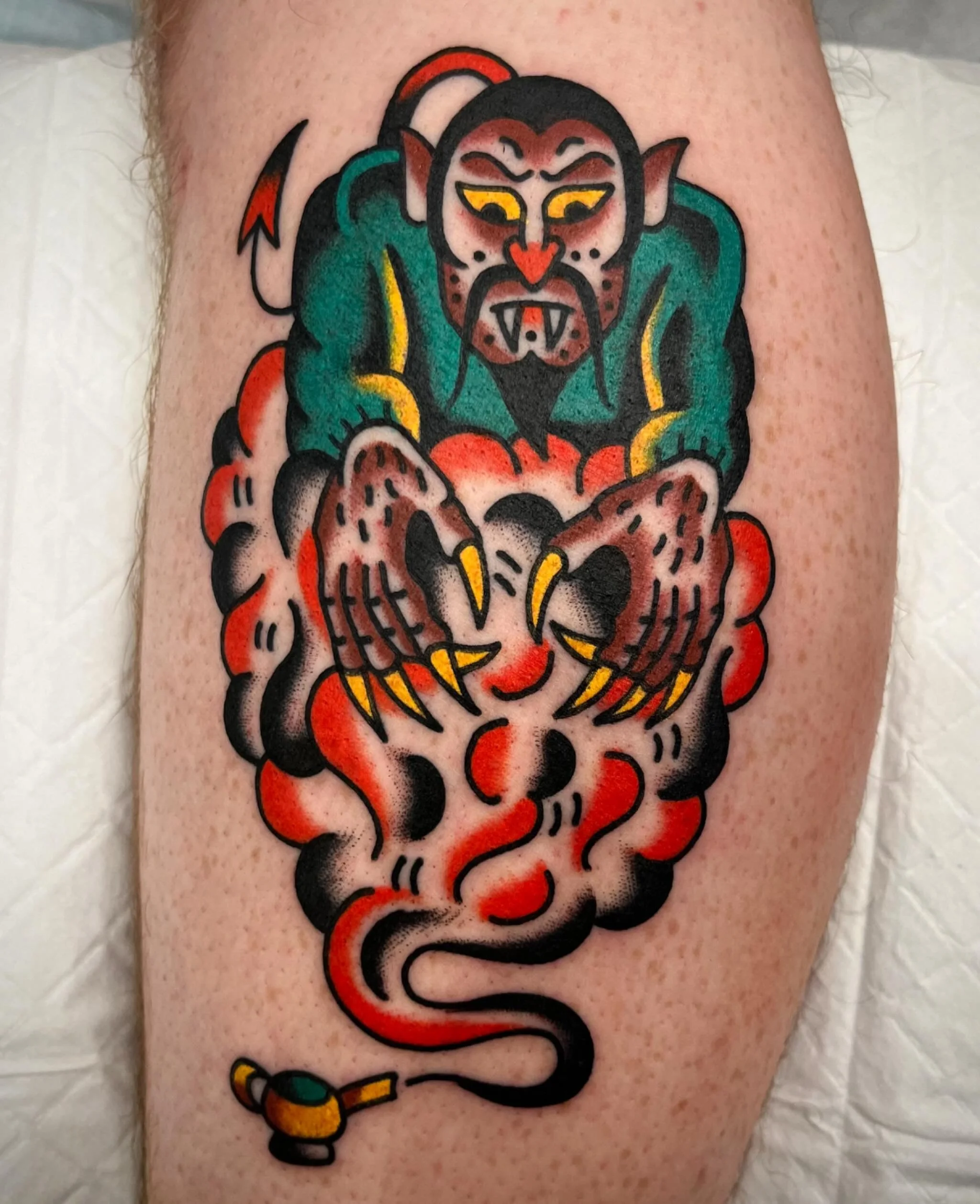 A tattoo of a traditional monster with green skin, red eyes, fangs, and a beard, holding a pig with claws, surrounded by smoke and clouds, and a small hammer at the bottom.