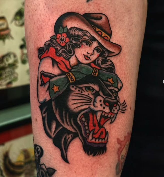 Tattoo of a woman with wavy hair and a flower in her hair, wearing a hat and a red scarf, combined with a fierce roaring black panther wearing a sheriff's deputy hat.
