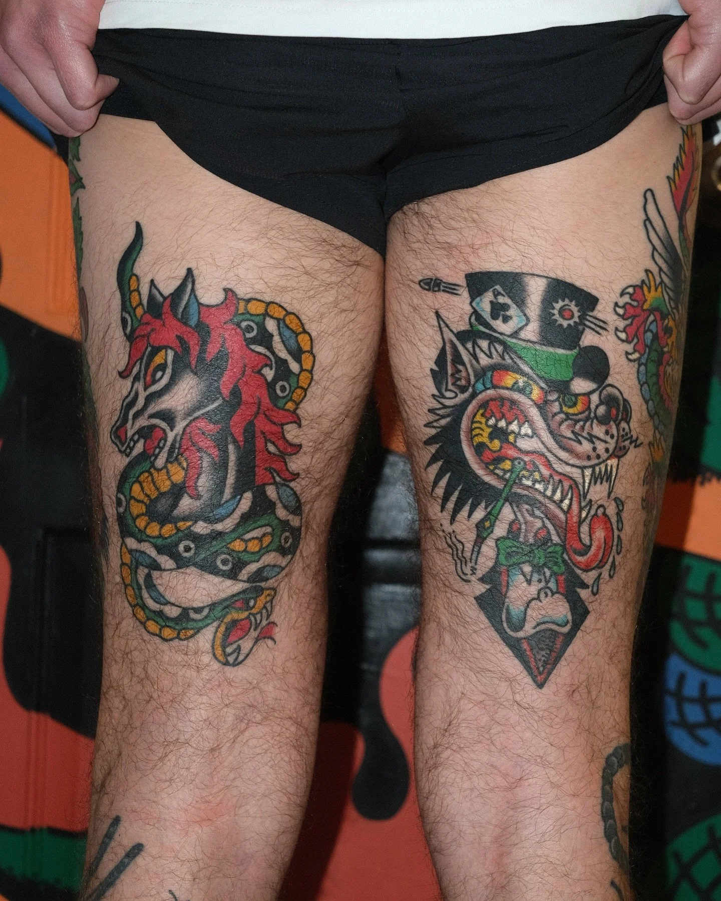Tattoo of a fierce tiger with sharp teeth and aggressive expression, wearing a ninja headband, on the right thigh. Tattoo of a colorful dragon with an open mouth and a fierce expression, also with sharp teeth, on the left thigh.