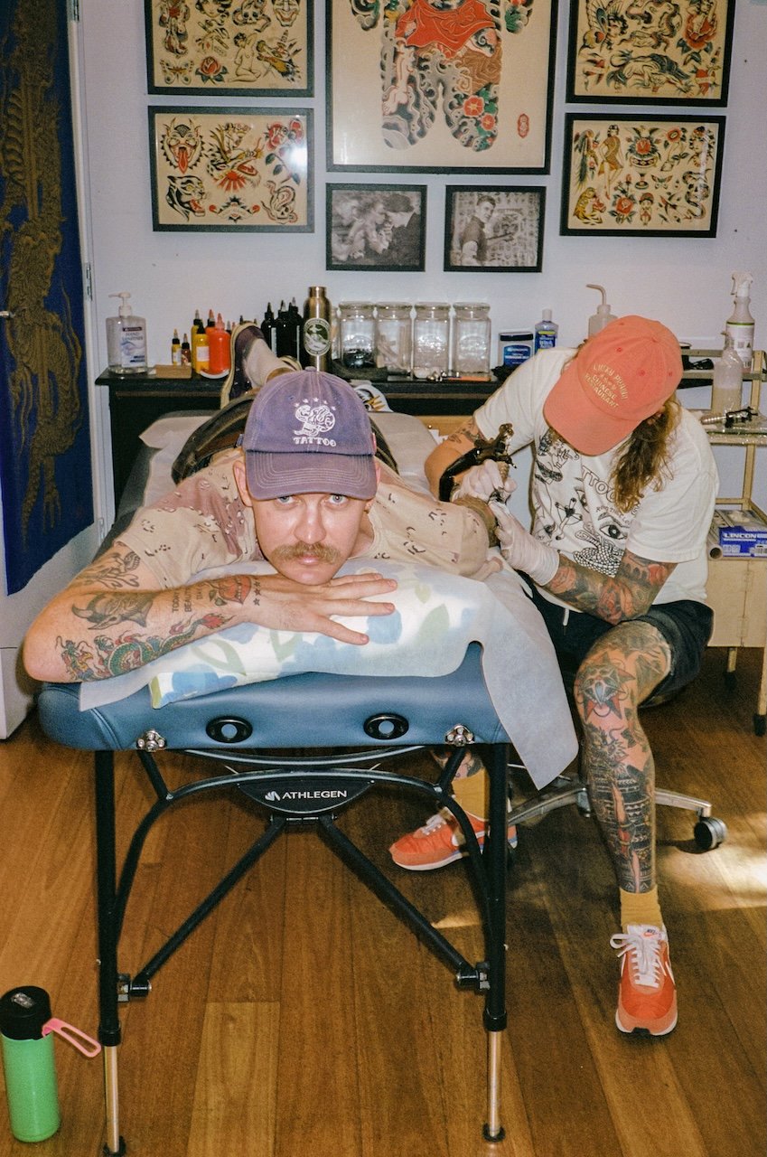 A man with tattoos and a mustache lying on a massage table, receiving a tattoo from a tattoo artist with tattoos on her arms in a tattoo studio. The man is wearing a purple cap, while the artist wears an orange cap and a white shirt. The studio has t