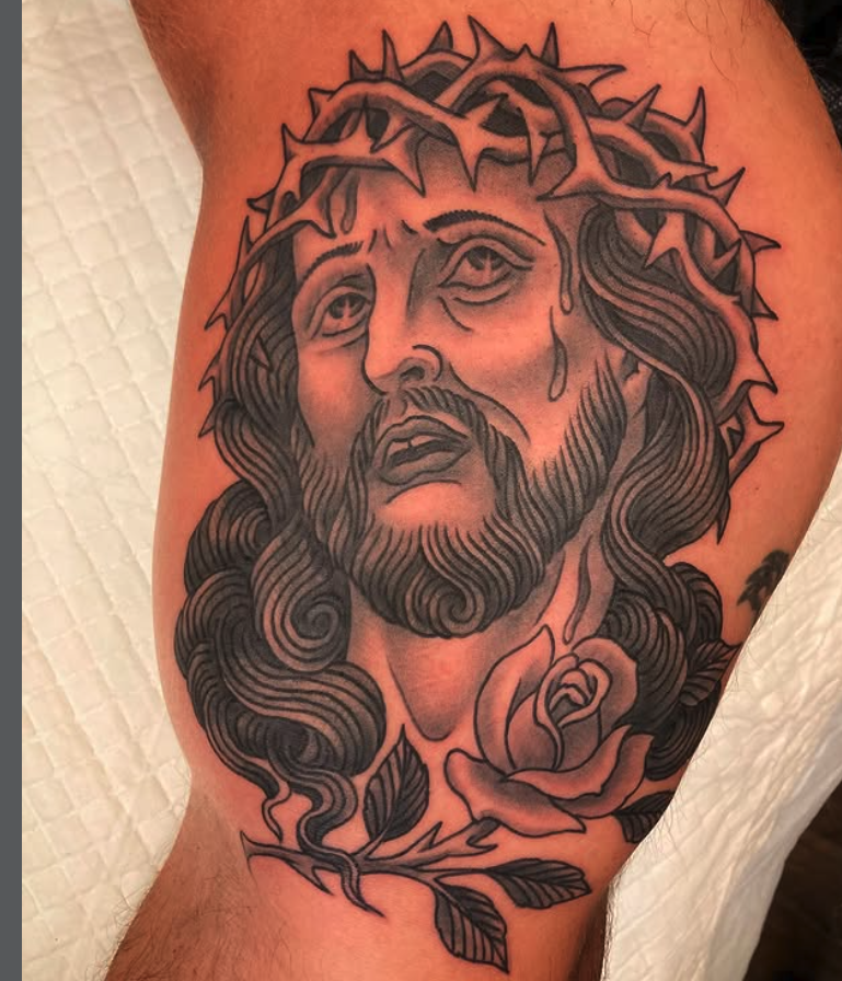 A tattoo of Jesus Christ wearing a crown of thorns, with long, wavy hair and a beard, surrounded by a rose and leaves.