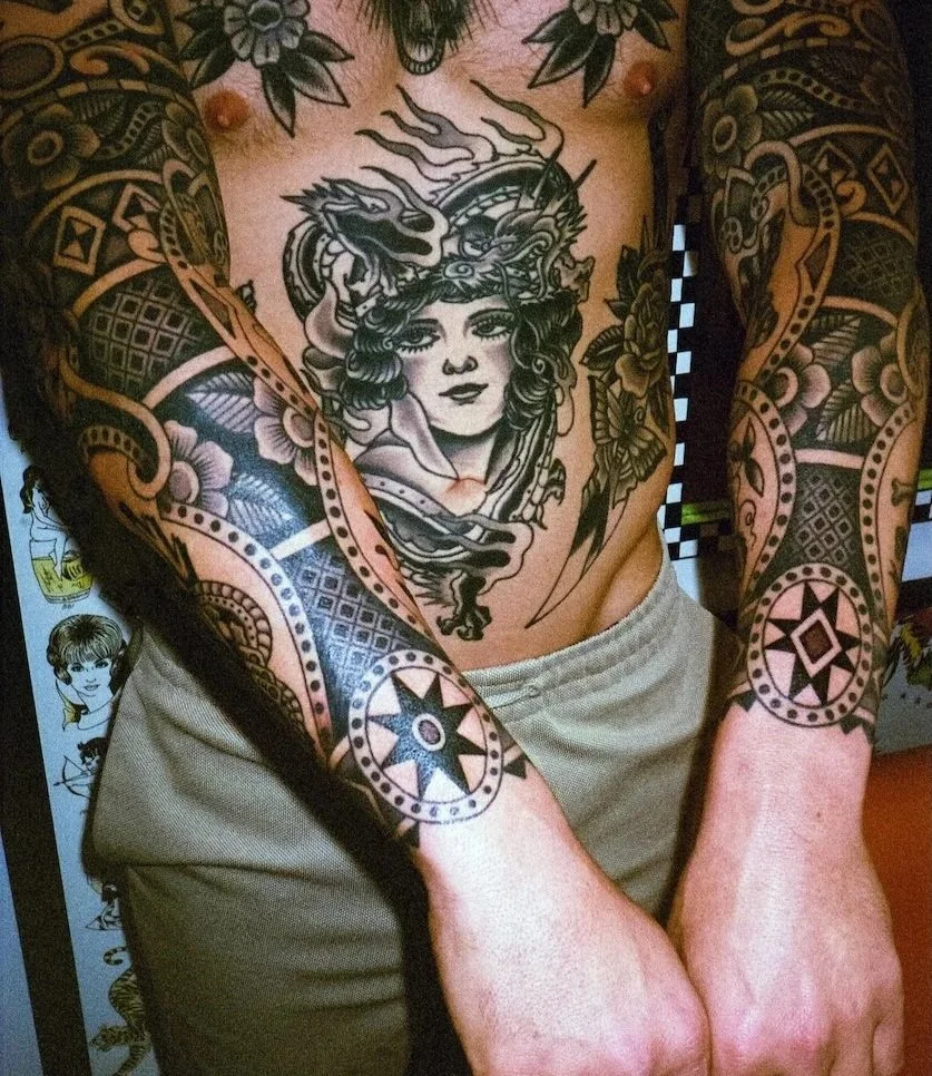 Close-up of a person's chest and arms covered in black and gray tattoos, including a large portrait of a woman with curly hair and a dragon on the chest, and various geometric and floral patterns on the arms.
