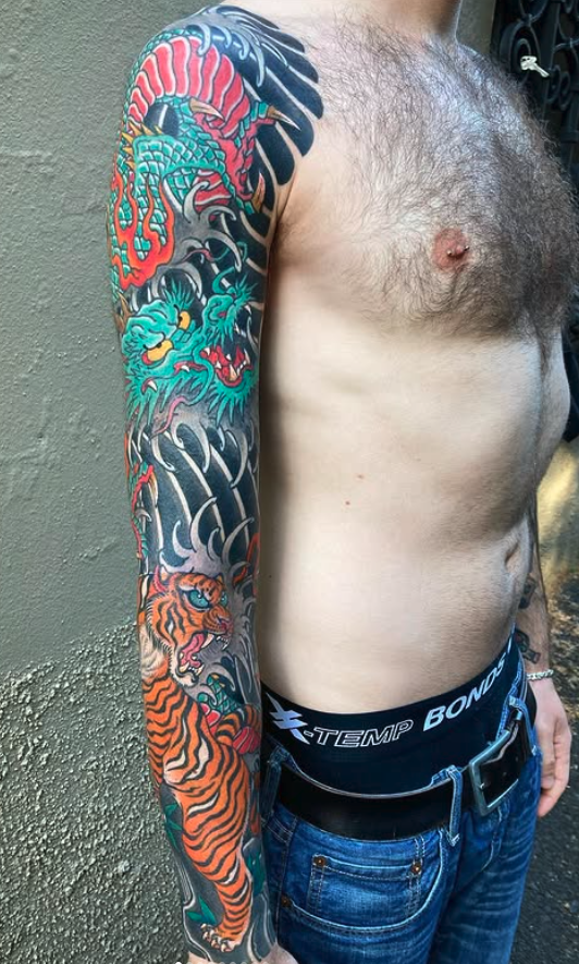 A person with a large, colorful sleeve tattoo depicting a dragon, a lion, and other mythical or animal elements on their left arm.