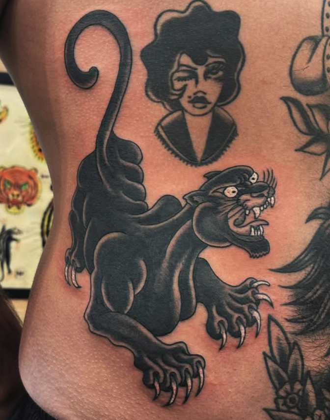 Tattoo of a ferocious black panther with open mouth, sharp teeth, and claws, beneath a portrait of a woman with wavy hair, in black ink.