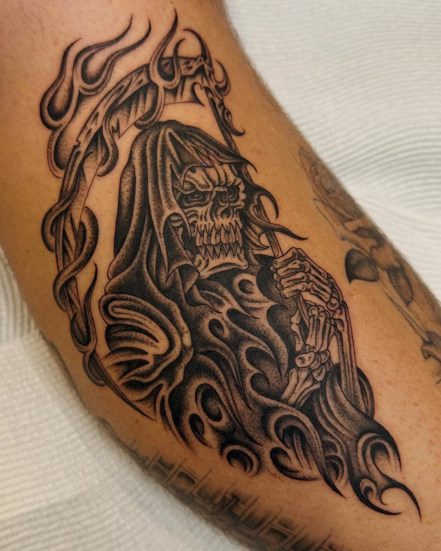 Black and gray tattoo of a skeletal figure with a skull face, hooded cloak, and long hair, holding a chain weapon, with flowing abstract elements surrounding it.