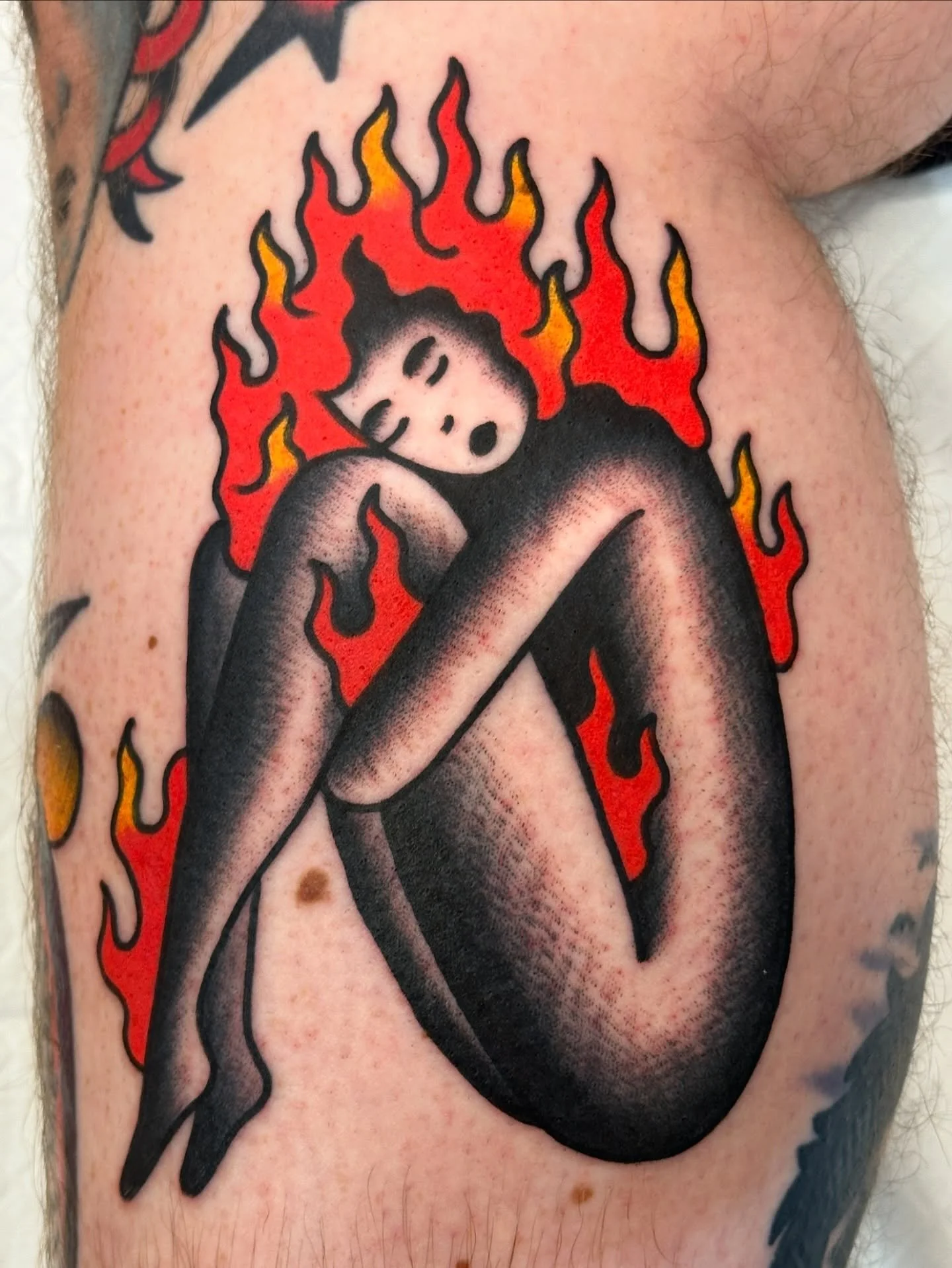 Tattoo of a stylized woman with black hair, closed eyes, and an open mouth, surrounded by red and yellow flames, with a black teardrop-shaped object near her face.