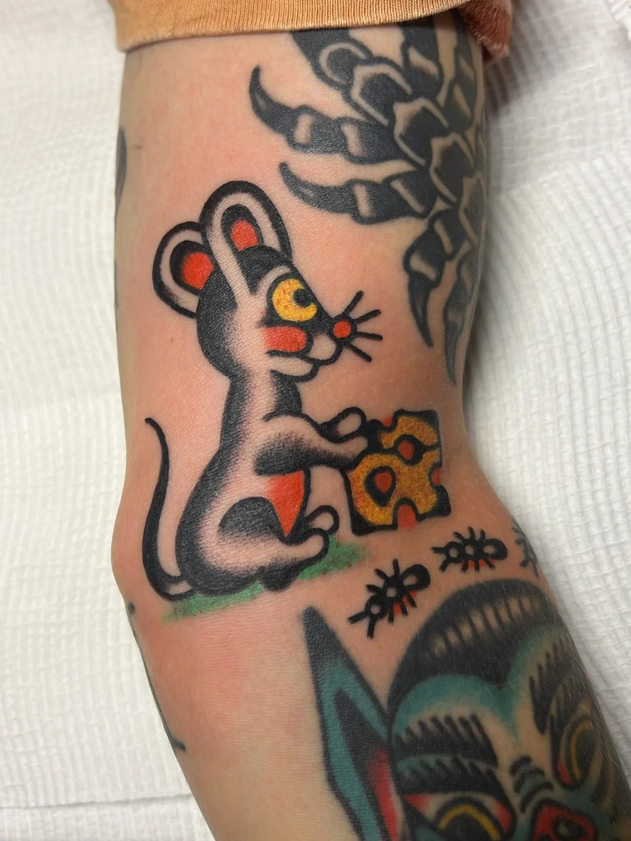 Tattoo of a cartoon-style mouse with yellow eye, sitting on grass with a piece of cheese, surrounded by other tattoo designs.