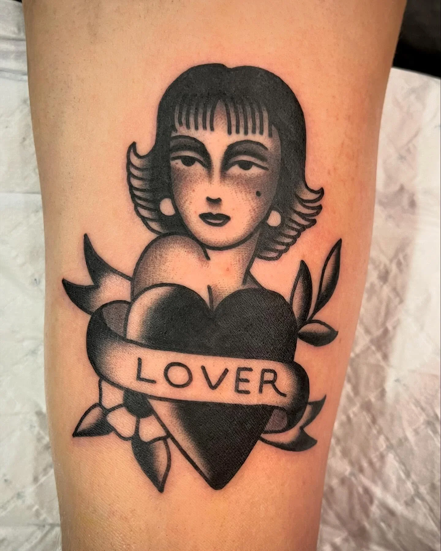 Tattoo of a woman with vintage hairstyle, a heart with a banner reading 'LOVER', surrounded by leaves and flowers.