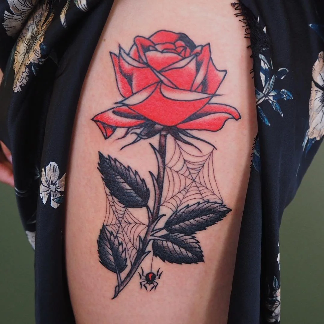 A tattoo of a red rose with black leaves, spider web details on the stem, and a small spider hanging from it.