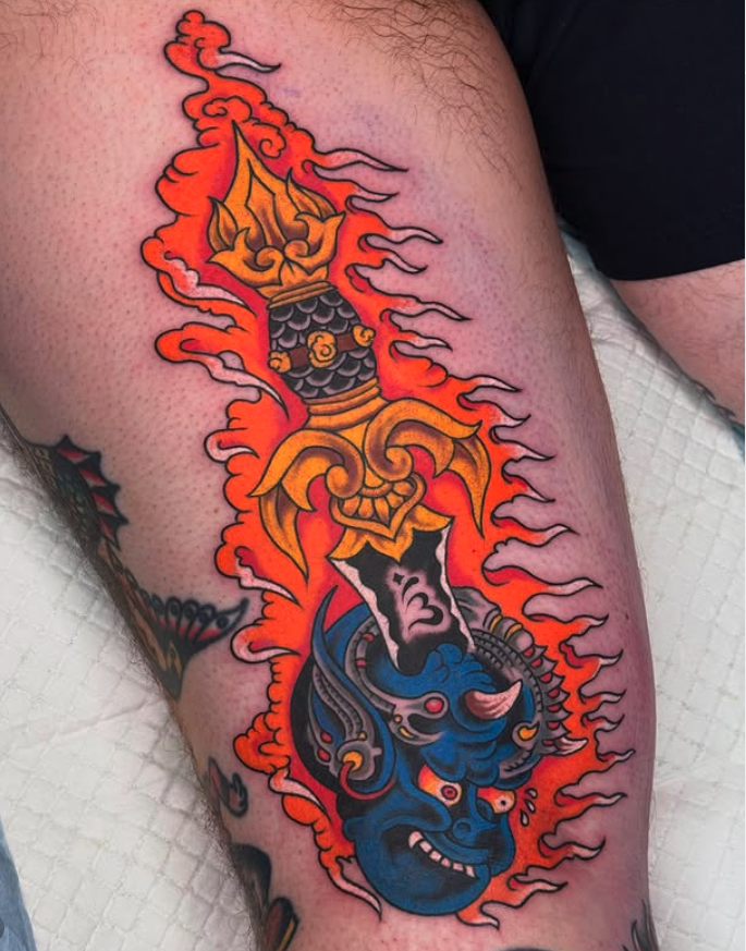 Colorful tattoo of a blue face with angry expression, surrounded by flames, with intricate patterns and a flaming sword above the face.
