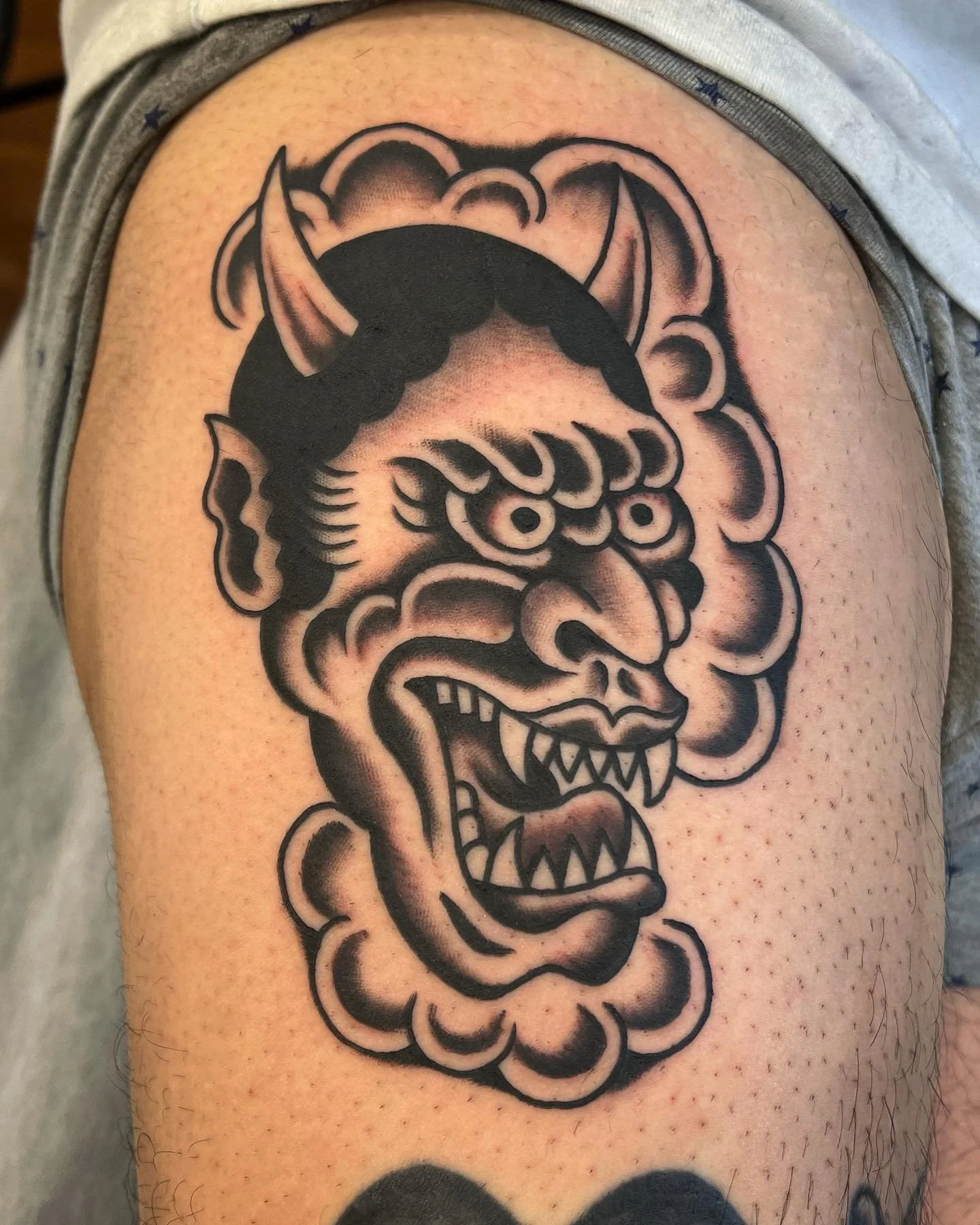Tattoo of an angry, traditional-style mask with sharp teeth, prominent nose, and horns on a person's upper arm.