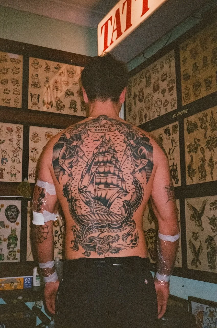 Back view of a man with tattoos, standing in front of walls filled with tattoo sketches, with a tattoo shop sign partially visible at the top.