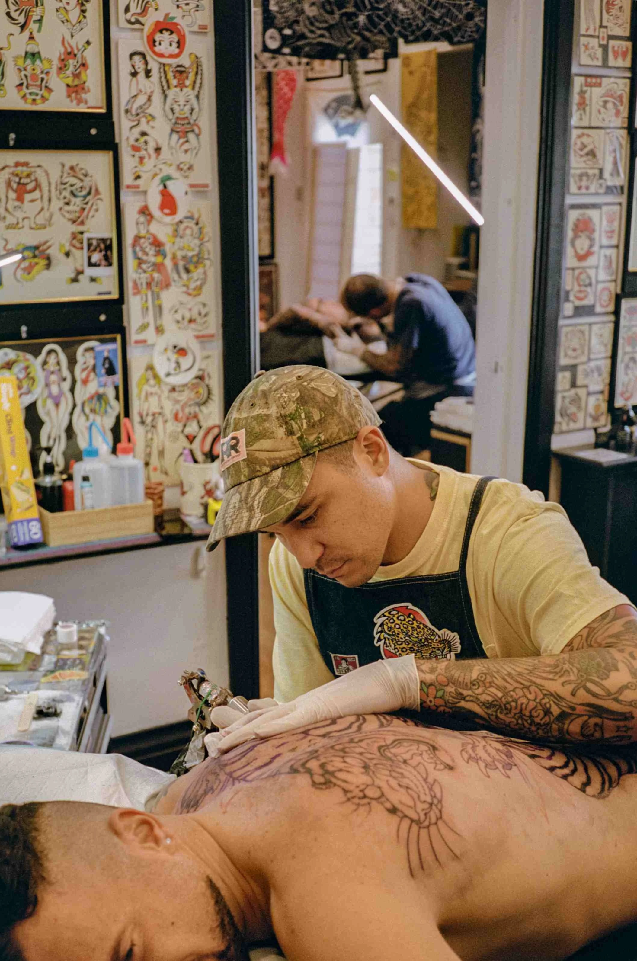 A tattoo artist working on a large tattoo on a man's back in a tattoo studio. The studio walls are decorated with tattoo flash and artwork. A person in the background is also getting a tattoo.