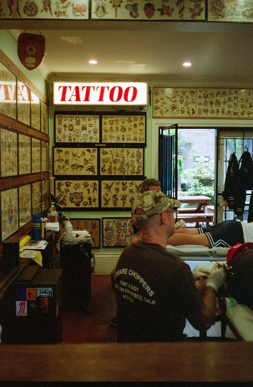 Inside a tattoo parlor with tattoo flash art on the walls, a bright 'TATTOO' sign, and a person receiving a tattoo while lying on a chair. The tattoo artist is wearing gloves and a T-shirt with 'SMARE CHOPPERS' printed on it.