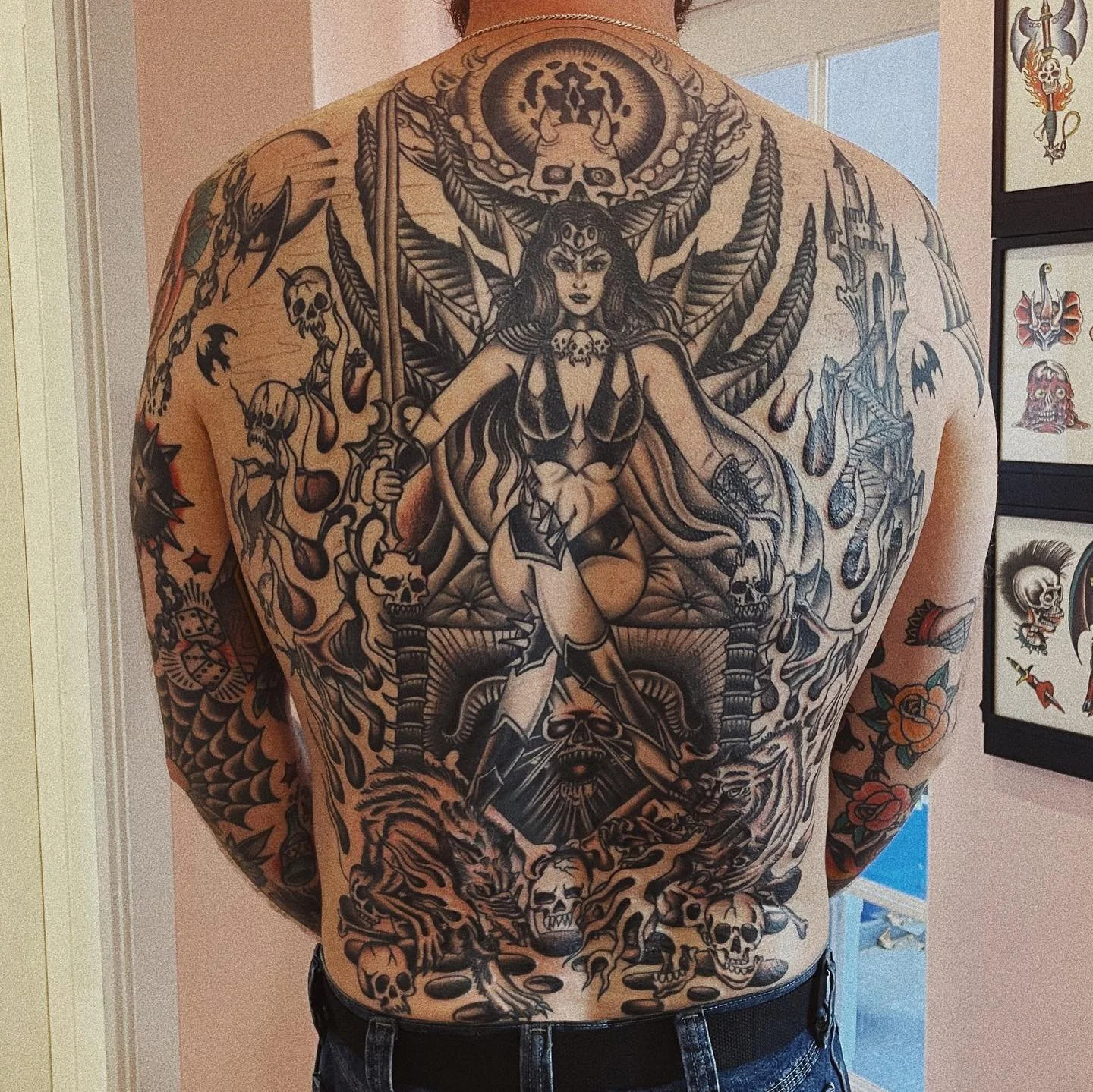 A man with a large, detailed black tattoo covering his back, featuring a woman holding a sword, surrounded by skulls, flames, and fantasy elements.