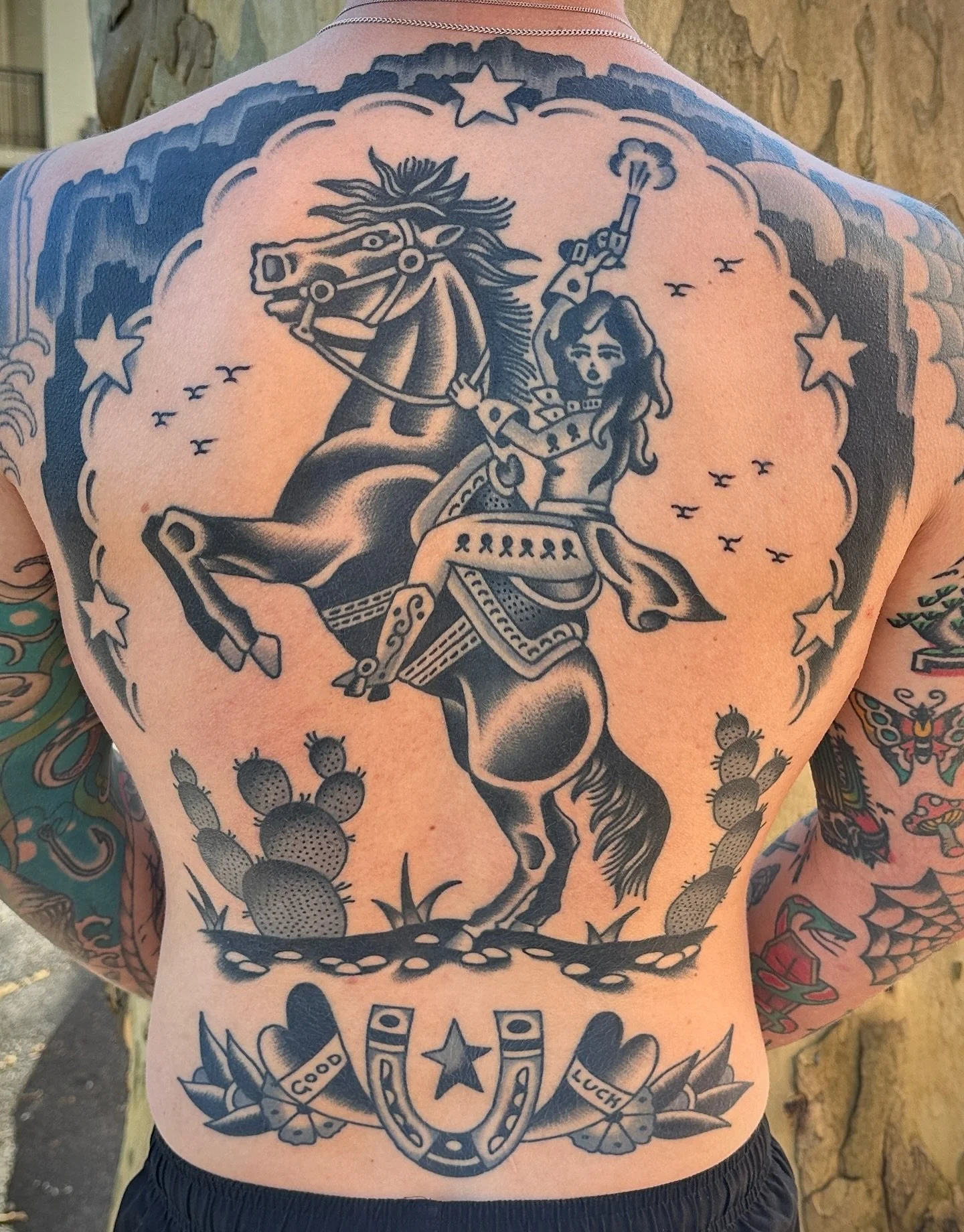A large back tattoo of a woman riding a rearing horse, holding a gun, surrounded by stars, cacti, and decorative elements, with the words 'Good' and 'Luck' in banners at the bottom.
