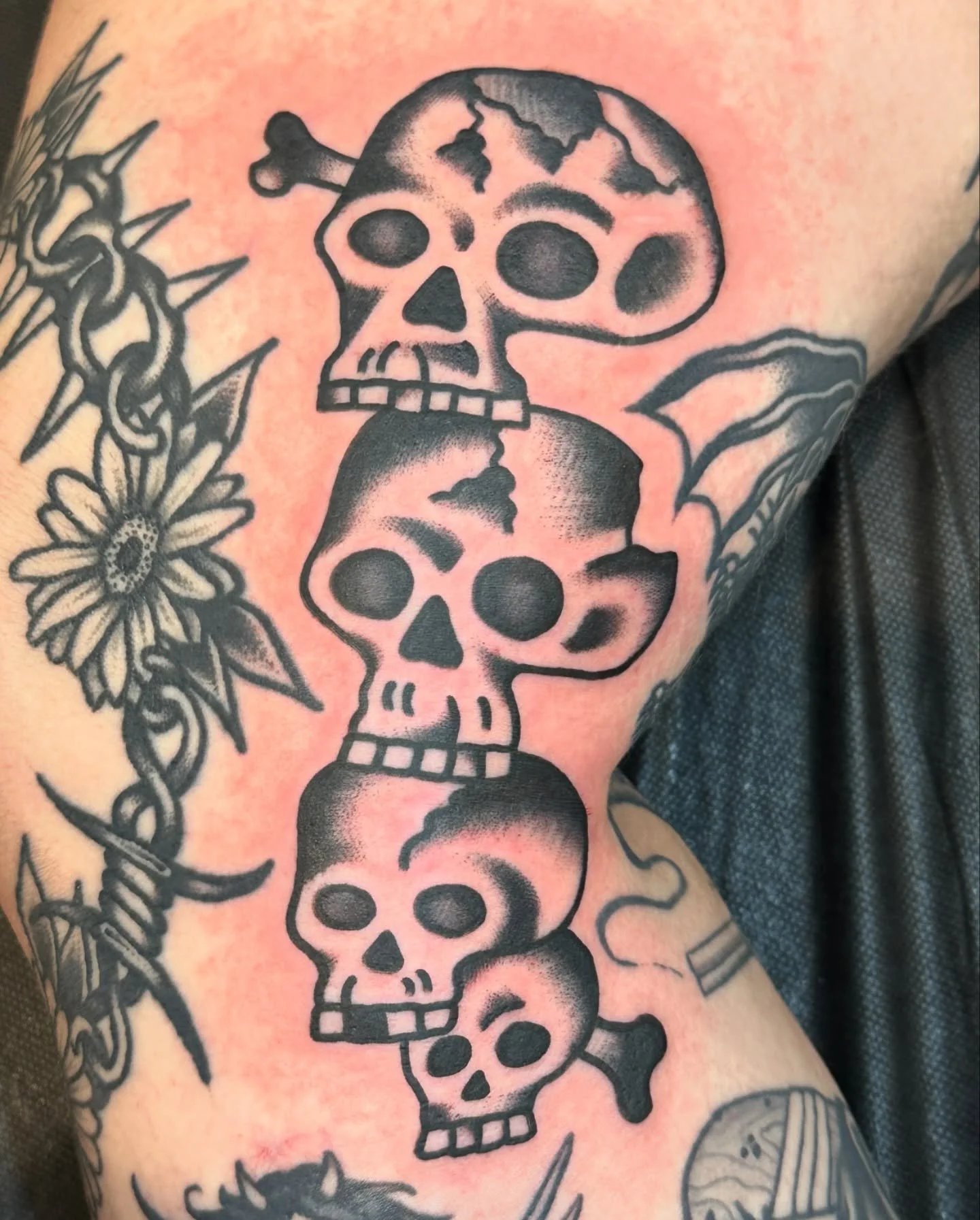 Tattoo of five black and white skulls arranged vertically, with some skulls having cracks and one skull with bat wings. Surrounding the skulls are additional tattoos, including a flower with a chain and other designs.