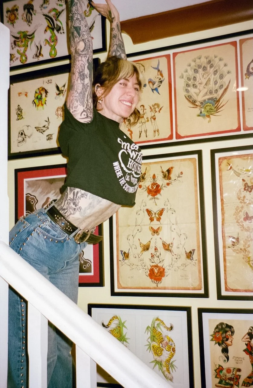A woman with tattoos on her arms and stomach, wearing a black t-shirt and jeans, is smiling and standing on a staircase with her arms raised above her head, in front of a wall decorated with framed tattoo artwork.