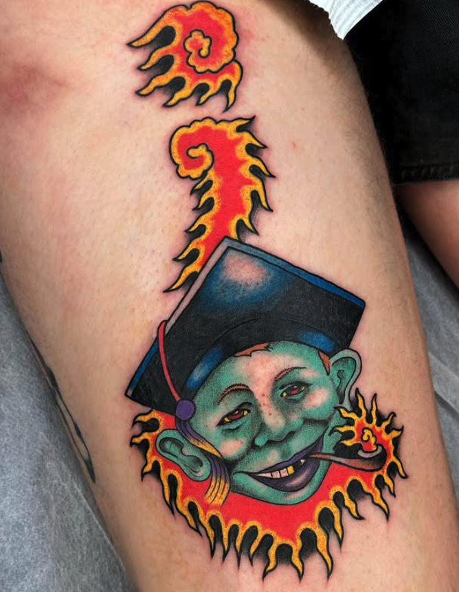 Tattoo of a smiling goblin wearing a graduation cap, smoking a pipe, surrounded by flames and a smoking flame design above.