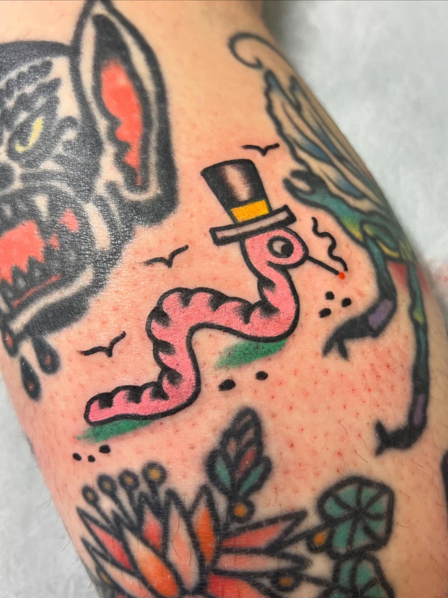 Close-up of a tattoo featuring a pink snake wearing a black top hat with a yellow band, with black birds flying around and other colorful floral and abstract elements visible.