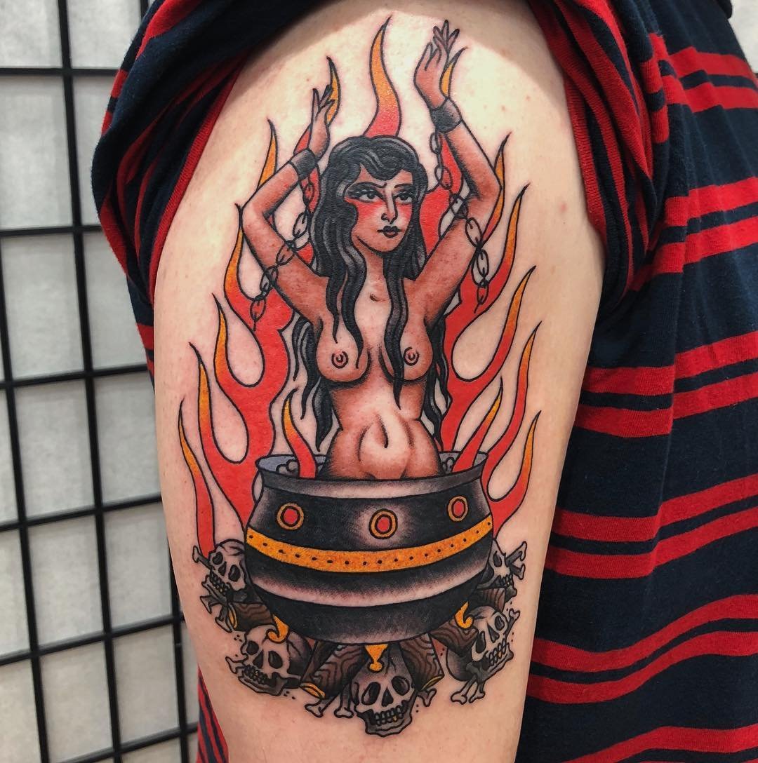 Tattoo of a woman with long hair, topless, with arms raised, surrounded by flames, standing inside a cauldron with skulls around its base.