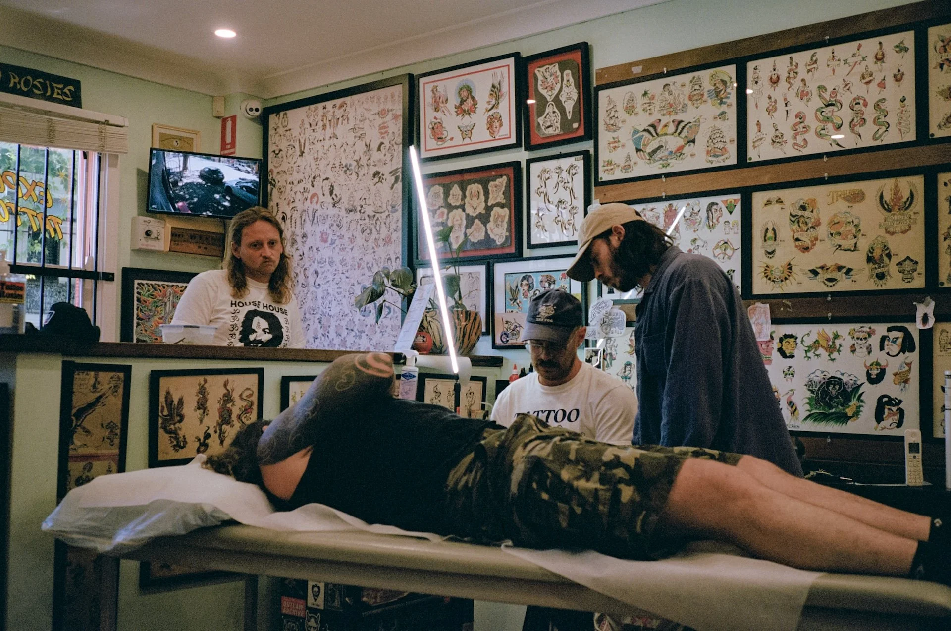 Person lying face down on a tattoo studio bed, surrounded by two tattoo artists and a customer, with tattoo flash art on the walls.