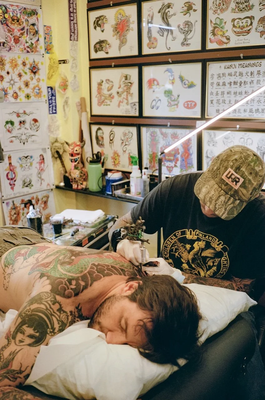 A tattoo artist working on a man's back in a tattoo shop, surrounded by tattoo designs on the walls.