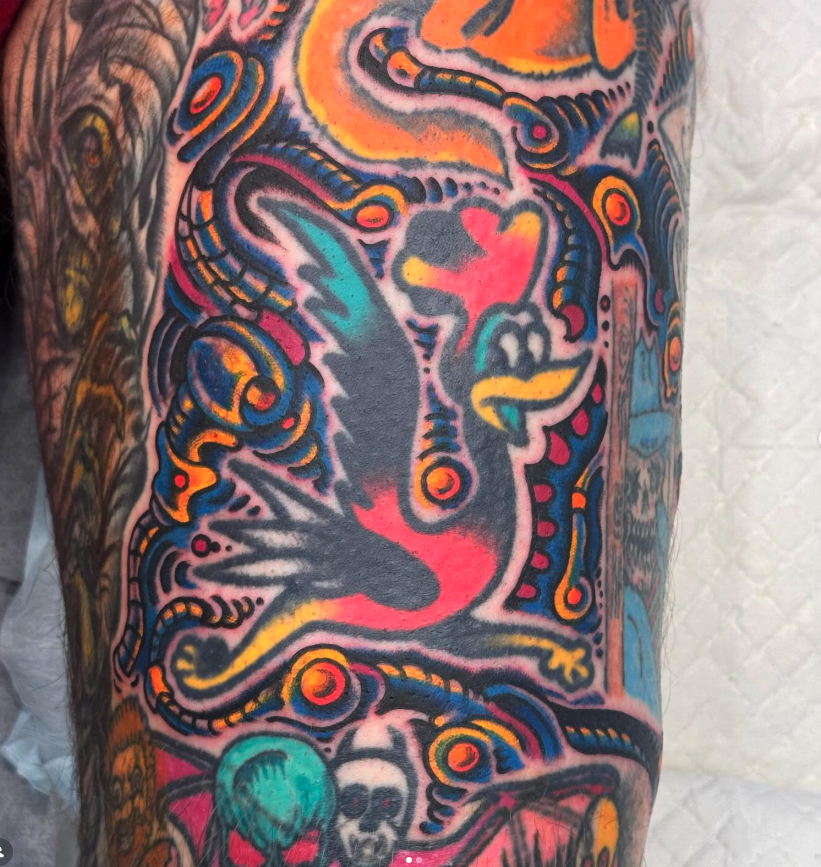 Bright and colorful tattoo featuring a stylized bird with outstretched wings, surrounded by abstract patterns, swirls, and skull images.