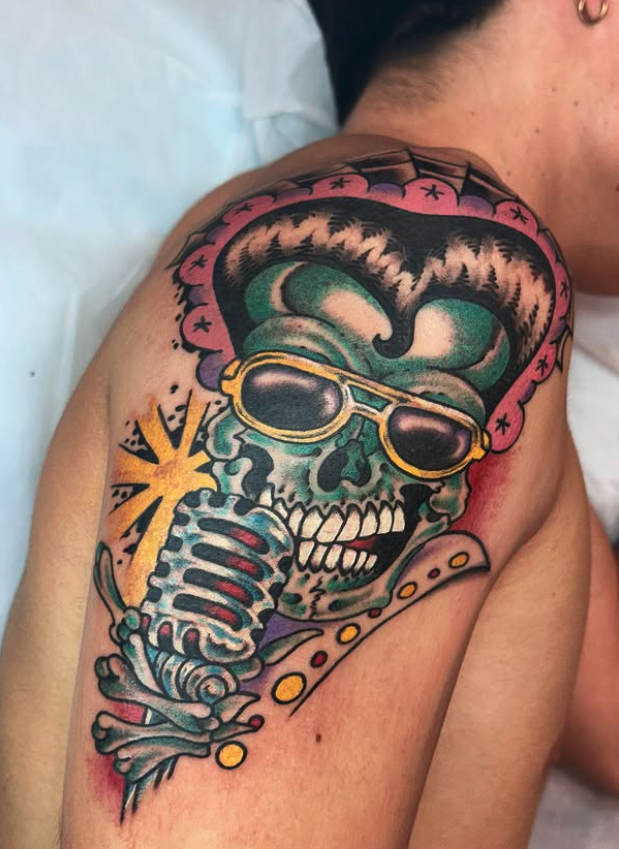 Tattoo of a skull with sunglasses, holding a vintage microphone, wearing a cowboy hat, with a yellow starburst behind the microphone.