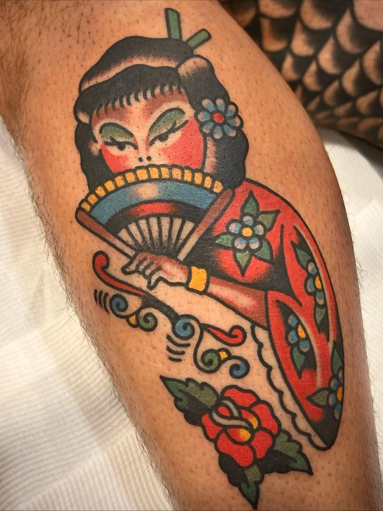 Tattoo of a traditional Asian geisha holding a folding fan, with floral and decorative details in red, blue, green, black, and yellow on the arm.