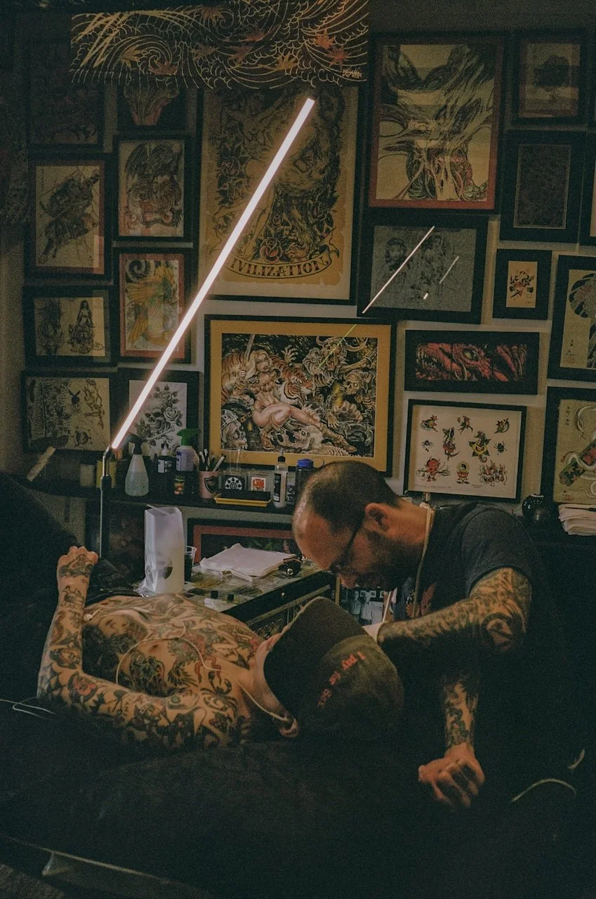 Tattoo artist working on a client's arm in a tattoo studio with tattoo flash art on the walls.