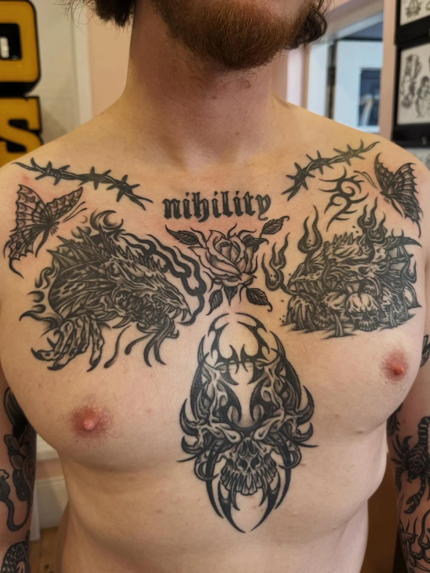 A man with a chest tattoo featuring a dragon, skull, and floral designs, with the word 'nobility' above the tattoos, barbed wire across the shoulders, and butterflies.