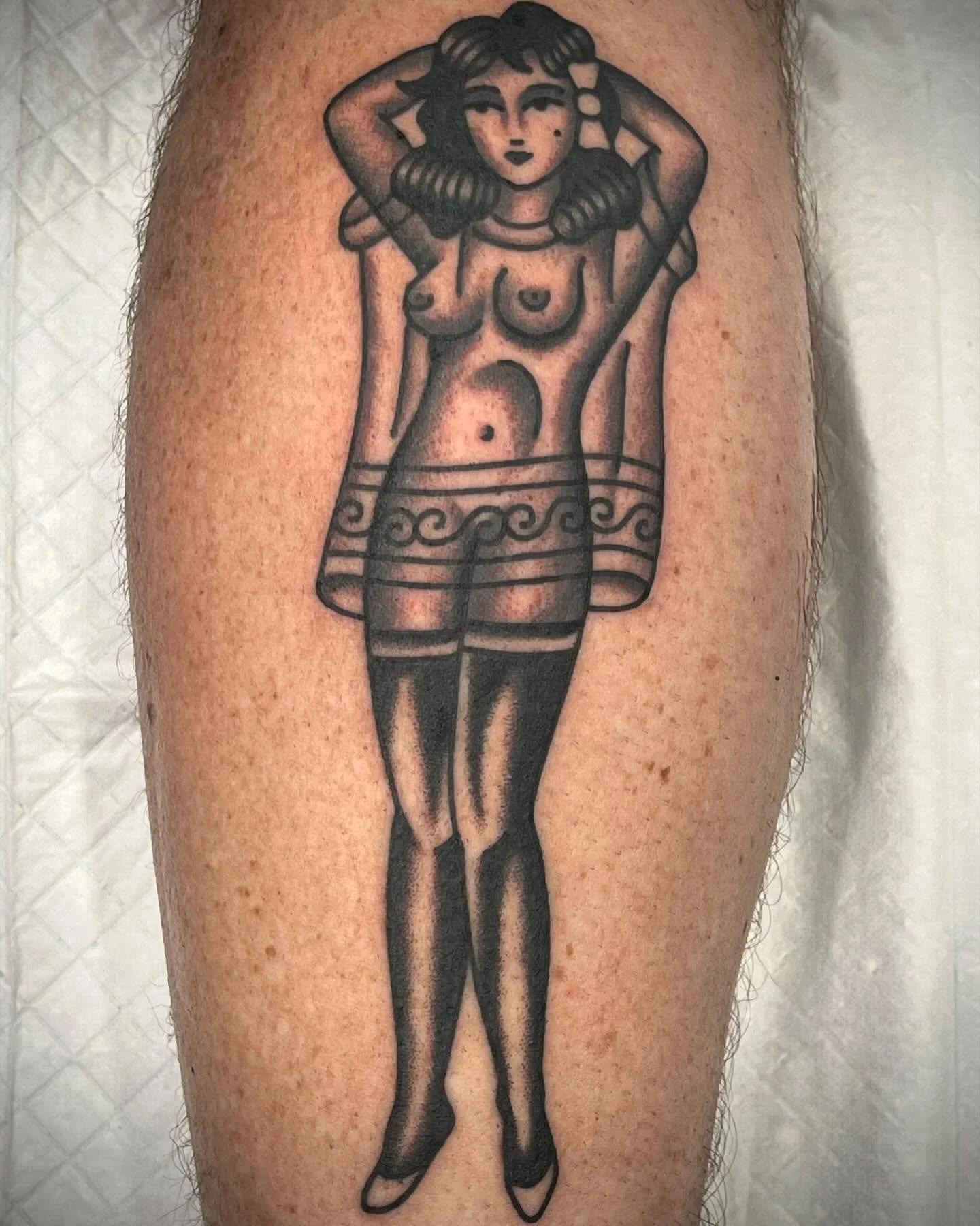 Tattoo of a woman with her arms behind her head, dressed in a vintage pinup style, wearing thigh-high stockings, and a decorative skirt.