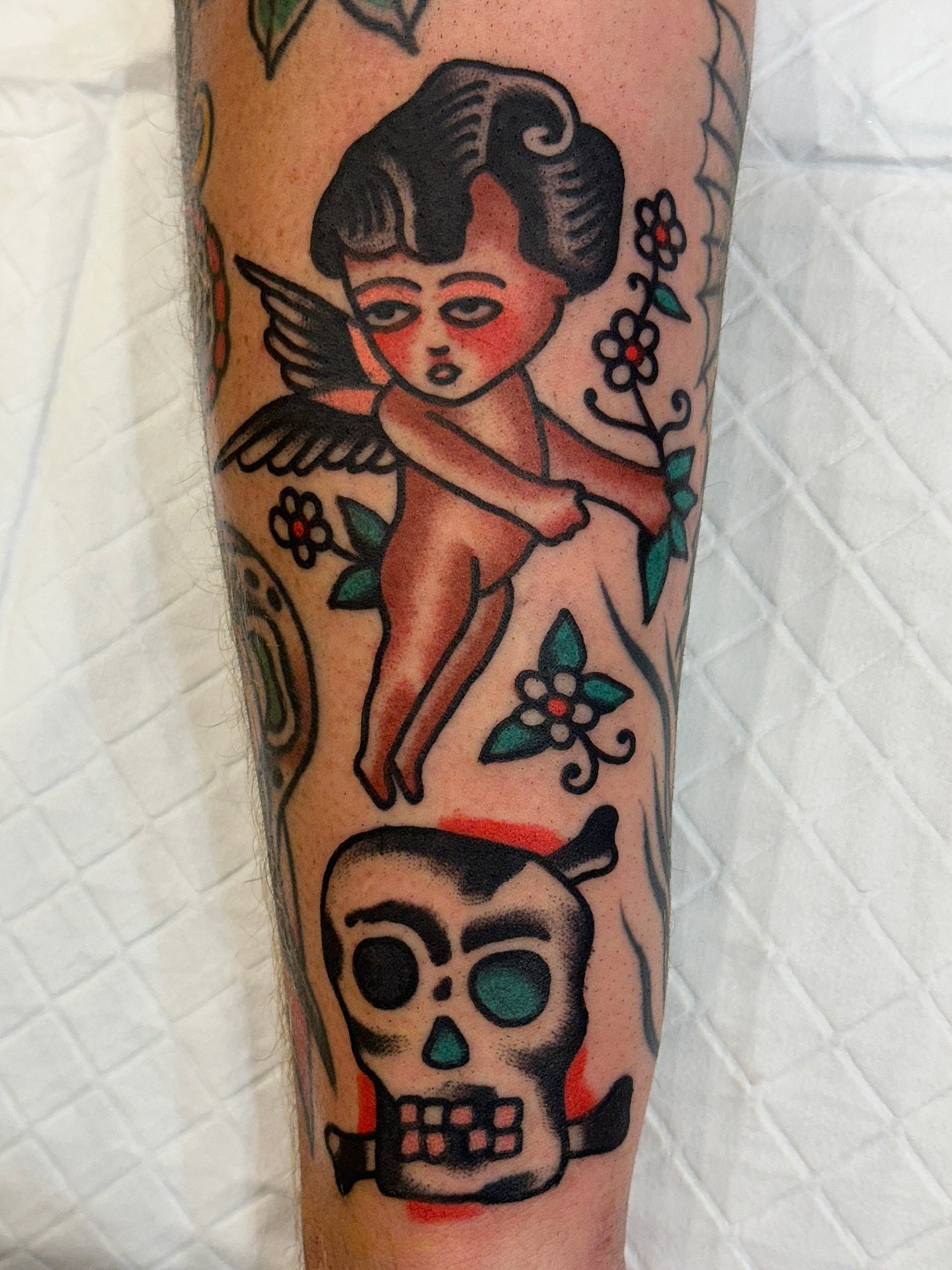 Tattoo of a cartoon Cupid with black hair, wings, and a bow, aiming an arrow at a skull with a cigar in its mouth, on a person's arm.