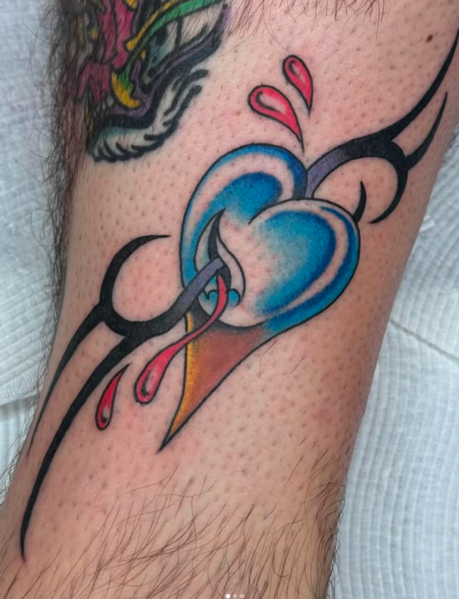 Colorful tattoo of a blue heart with black outlines and pink drips, surrounded by black line designs and pink accents on skin.