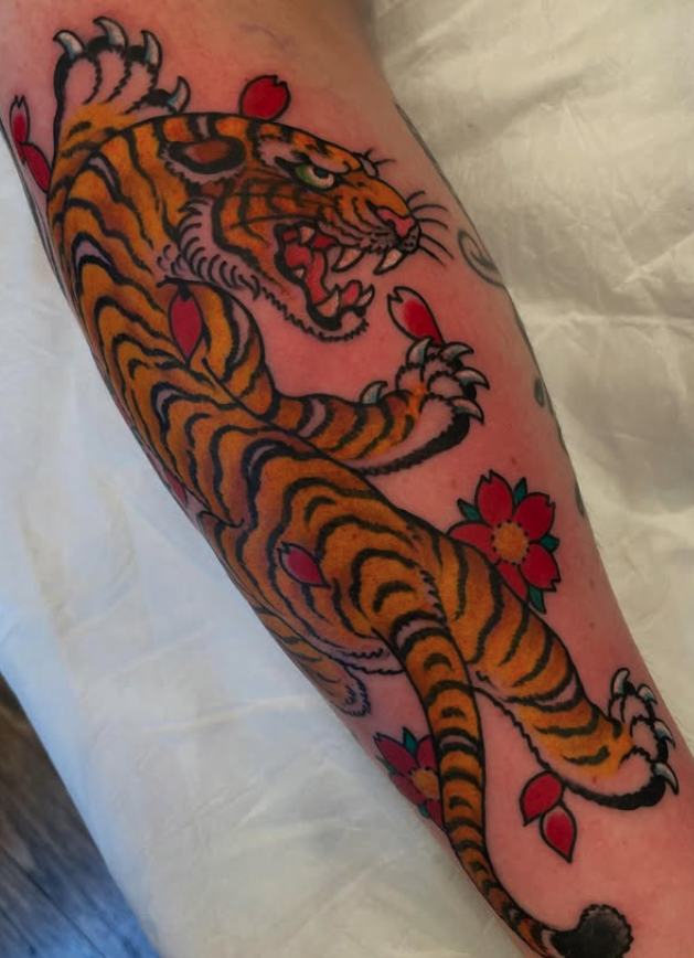 Tattoo of a roaring tiger with black stripes, surrounded by pink and red flowers on a person's arm.