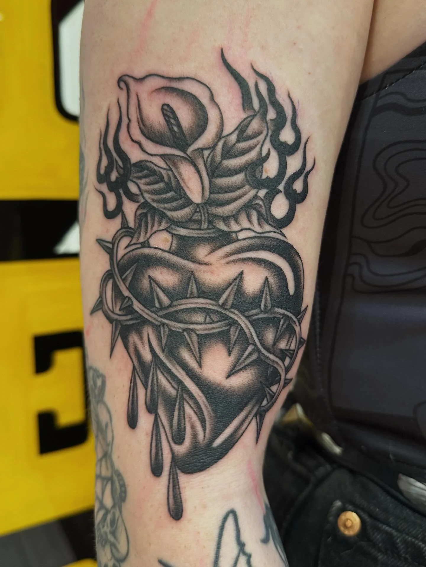 Black and gray tattoo of a heart pierced by a thorny vine, with flames and a rose emerging from a cracked skull in a dark, illustrative style.