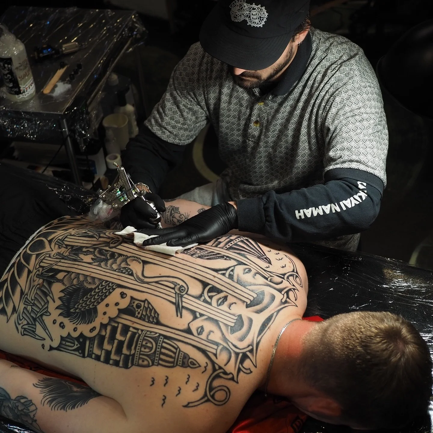 Tattoo artist in black gloves is tattooing a large black and gray design on a person's back, who is lying face down on a tattoo chair. The artist wears a baseball cap and a long-sleeve shirt with Asian-inspired patterns and text on the sleeve. The workspace includes tattoo equipment, ink bottles, and other supplies.