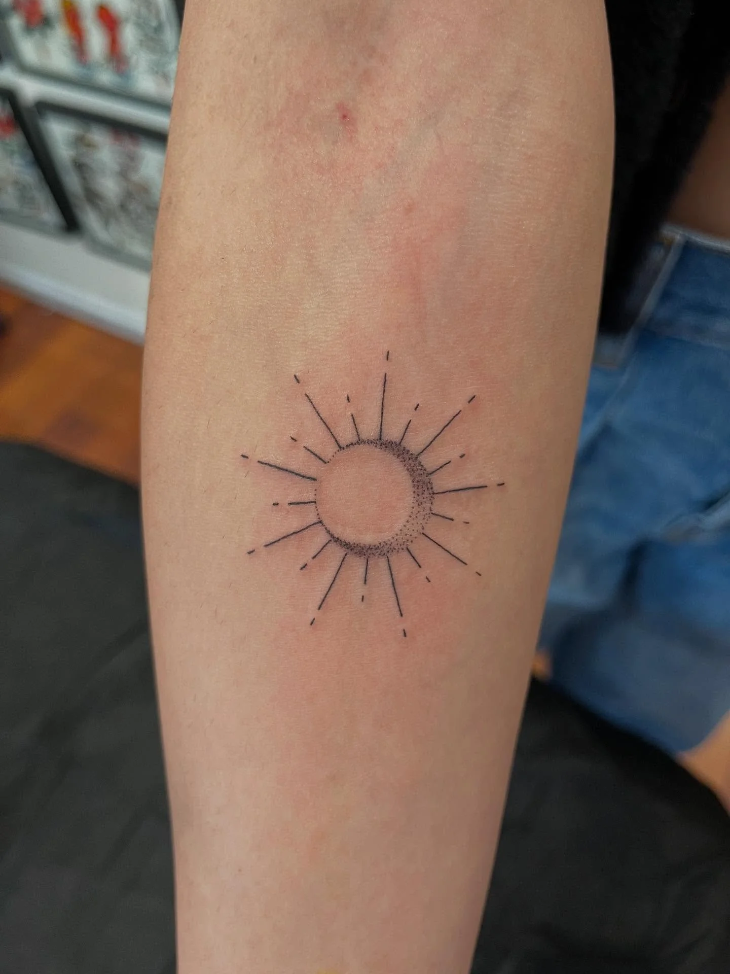 Tattoo of a crescent moon surrounded by lines representing the sun on a person's forearm.