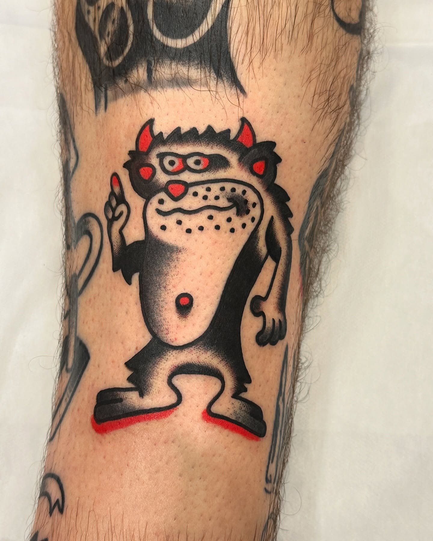 Tattoo of a cartoon-style creature with a humanoid body, three eyes, and devil-like horns. The creature is making a gesture with its right hand, and its tongue is sticking out.