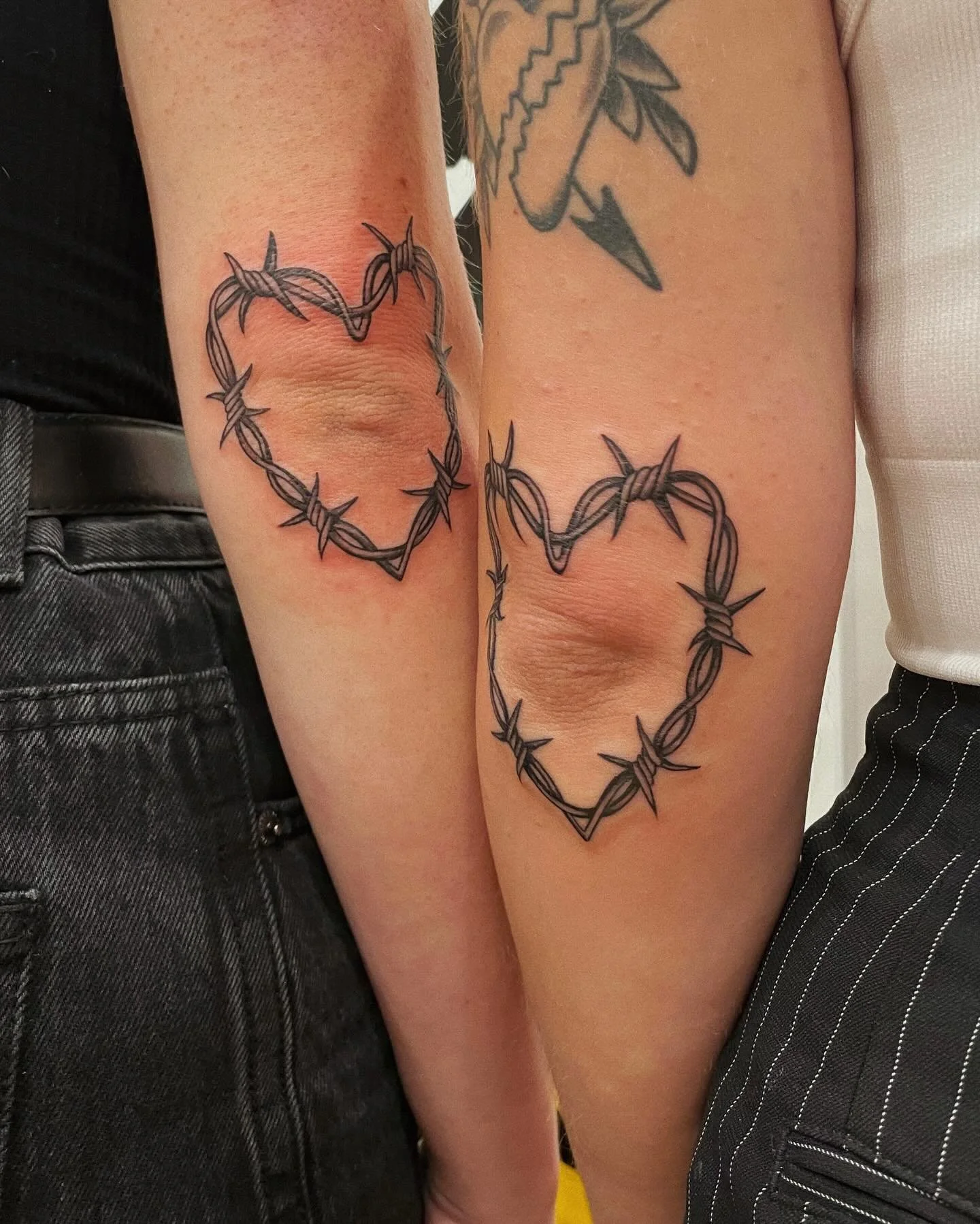 Tattoo of two intertwined hearts made of barbed wire on two people's arms.