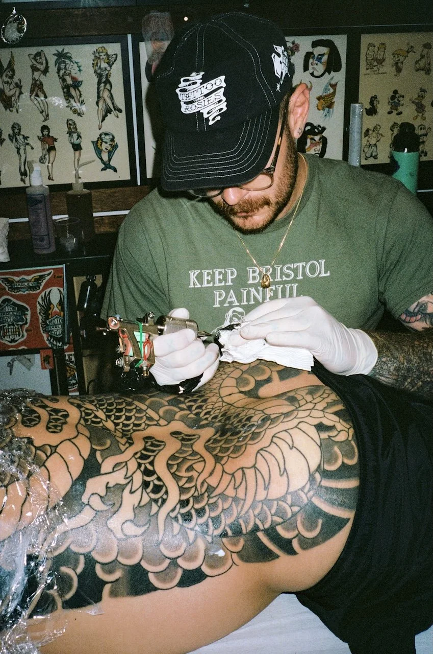 Tattoo artist working on a large, detailed dragon tattoo on a person's leg in a tattoo studio.